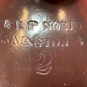 Pode incluir: Close-up de um jarro de cerâmica castanho com texto em relevo. O texto diz "A & L P NORTON, BENNINGTON, VB" e o número "2" também está em relevo. O jarro tem um acabamento brilhante e é provavelmente vintage.