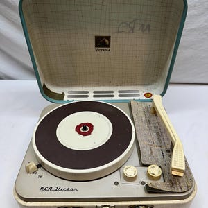 May include: A portable RCA Victor record player in a cream-colored case with a teal handle and trim. The interior lid features a grid pattern and a Victrola logo. The turntable is brown and white with a cream-colored tonearm and control knobs.
