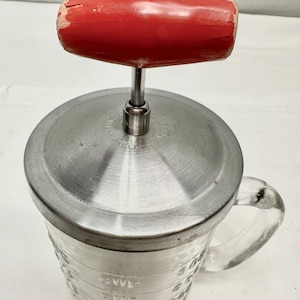 May include: A vintage glass measuring cup with a stainless steel lid and a red wooden handle. The cup has markings for measuring in ounces and cups.