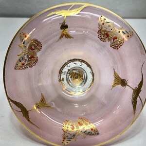 May include: A pink glass bowl with gold trim and butterfly and floral designs. The butterflies have red, blue, and gold accents. The bowl's rim and base are trimmed in gold.