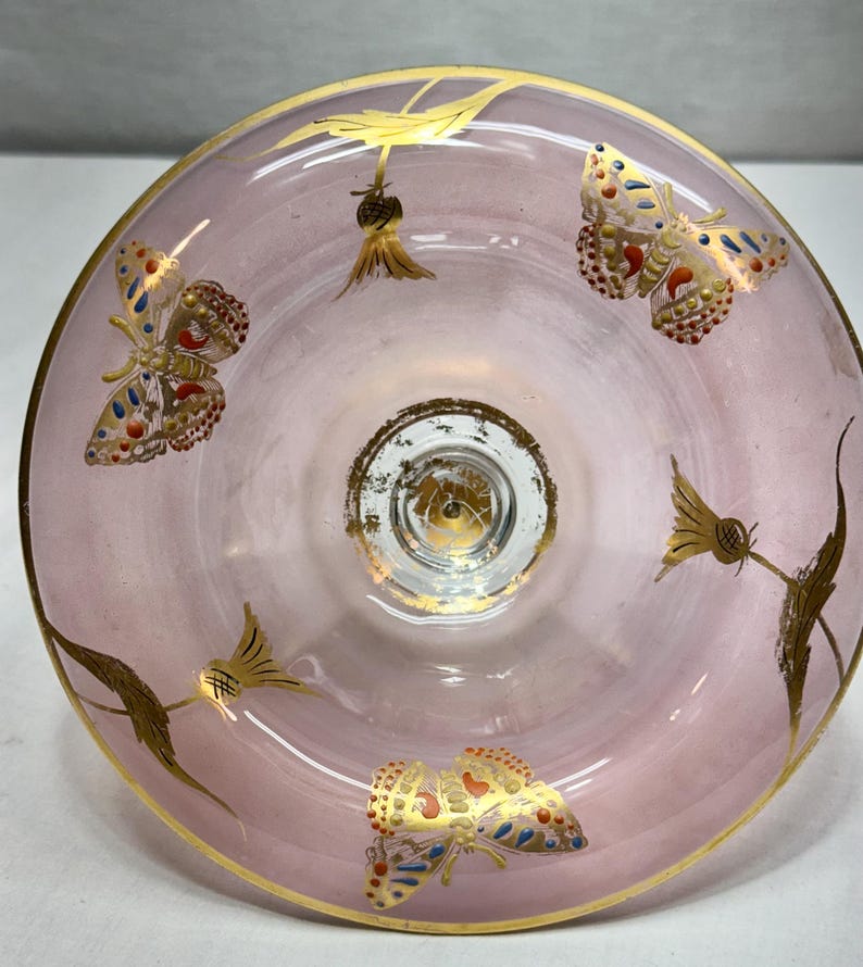 May include: A pink glass bowl with gold trim and butterfly and floral designs. The butterflies have red, blue, and gold accents. The bowl's rim and base are trimmed in gold.