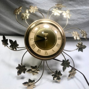 May include: Gold-toned wall clock with a round face and Roman numeral markings. The clock is surrounded by a decorative design of gold-colored metal leaves.
