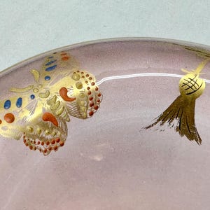 May include: Close-up of a decorative glass object with a pinkish hue. The object features gold painted butterfly and tassel designs with red, blue, and gold accents. The design is delicate and ornate.