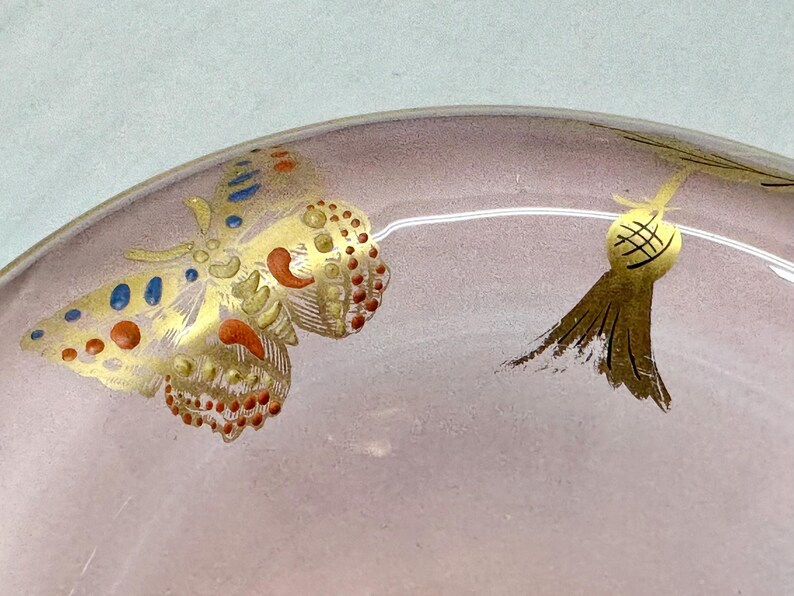 May include: Close-up of a decorative glass object with a pinkish hue. The object features gold painted butterfly and tassel designs with red, blue, and gold accents. The design is delicate and ornate.