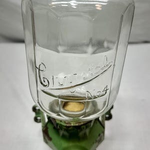 Op de afbeelding: Een doorzichtige glazen dispenser met een lichtgroen deksel en een groene metalen voet met een tuit. Het glazen lichaam is voorzien van de in reliëf gedrukte tekst Crystal No 4. De metalen voet vertoont slijtage en afbladderende verf.