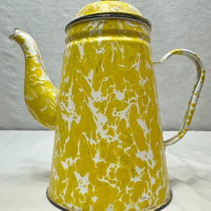 Könnte beinhalten: Vintage Kaffeekanne aus gelbem und weißem Emaille. Die Kanne hat einen Deckel, einen Ausguss und einen Griff. Das Design zeigt ein wirbelndes Muster aus Gelb und Weiß, was ihr ein Retro-Aussehen verleiht. Die Kanne ist wahrscheinlich aus Metall.