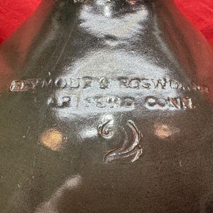 Pode incluir: Jarro de grés verde escuro com texto em relevo: "PLYMOUTH & ROSWOLD, AP 133RD CONN". O jarro tem um acabamento brilhante e uma pega curva. Um símbolo de lua crescente em relevo.