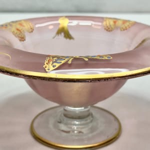 May include: A pink glass pedestal bowl with gold trim and butterfly and leaf designs. The bowl has a wide, shallow rim and a round base. The butterfly and leaf designs are in gold, blue, and orange. The bowl is likely used for serving desserts or displaying decorative items.