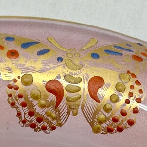 May include: A close-up of a pink glass object with a gold butterfly design. The butterfly has red, yellow, and blue accents. The wings are detailed with small dots and curved shapes.