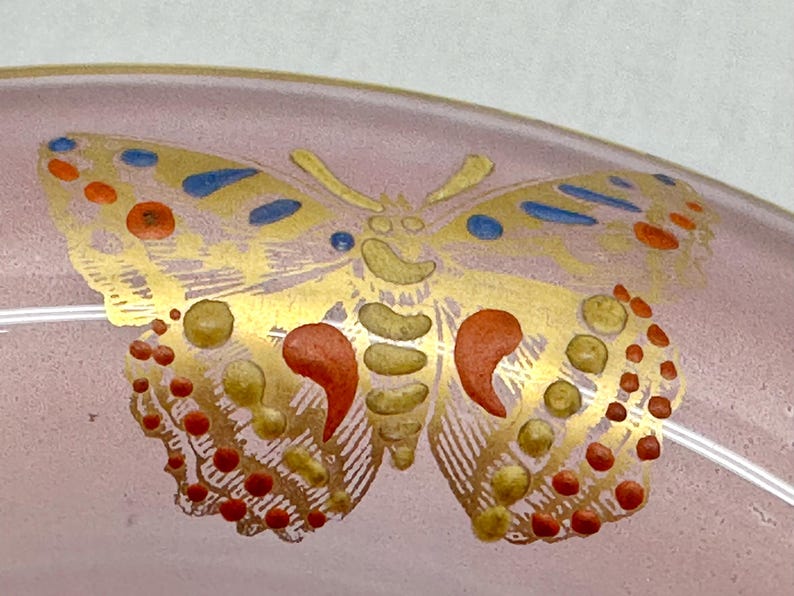 May include: A close-up of a pink glass object with a gold butterfly design. The butterfly has red, yellow, and blue accents. The wings are detailed with small dots and curved shapes.