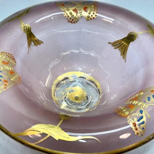 May include: A decorative, light purple glass bowl with gold trim and butterfly and leaf designs. The butterflies have blue, orange, and gold accents. The bowl has a clear, circular base.