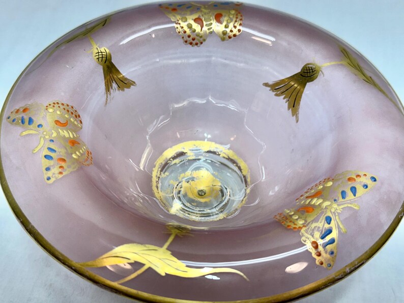 May include: A decorative, light purple glass bowl with gold trim and butterfly and leaf designs. The butterflies have blue, orange, and gold accents. The bowl has a clear, circular base.