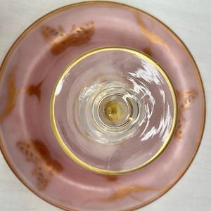 May include: Overhead view of a pink glass dessert plate and a clear glass with a gold rim. The plate has gold butterfly and swirl designs. The glass has a gold rim and a clear stem.