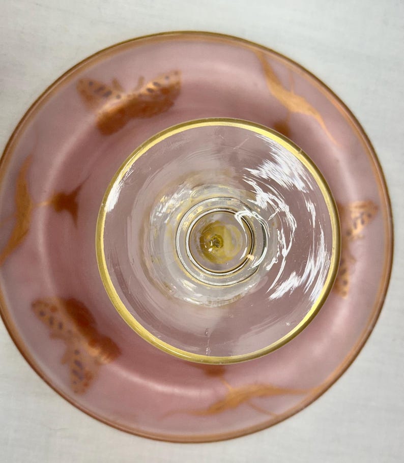 May include: Overhead view of a pink glass dessert plate and a clear glass with a gold rim. The plate has gold butterfly and swirl designs. The glass has a gold rim and a clear stem.