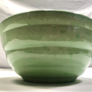 Puede incluir: Un cuenco de cerámica verde claro con un acabado brillante. El cuenco tiene una forma redondeada con estrías horizontales alrededor del exterior. El color pasa de un verde más oscuro en la base a un verde más claro hacia la parte superior. El cuenco es probablemente para servir comida.