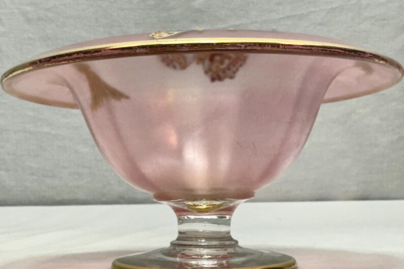 May include: A pink glass bowl with a flared rim and a pedestal base. The rim and base are trimmed with gold. The bowl has a delicate, vintage aesthetic, suitable for serving desserts or displaying small items.