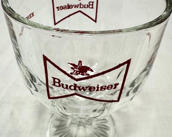 May include: A clear glass Budweiser beer goblet with a red Budweiser logo and an eagle design. The goblet has a fluted design and a stemmed base.