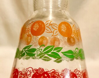 May include: A clear glass pitcher with a textured neck and a wide base. The pitcher is decorated with a repeating pattern of orange slices, green leaves, and red tomatoes.