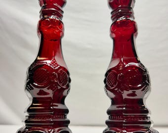 May include: A pair of deep red glass candle holders featuring an intricate pressed pattern with geometric shapes and circular motifs. Each holder has a hexagonal base, a tapered stem, and a wide top rim, showcasing a reflective, translucent finish.