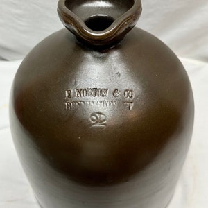 Pode incluir: Uma jarra de grés castanho com uma pega e um bico. A jarra é marcada com o texto "E Norton & Co. Bennington VT" e o número "2".