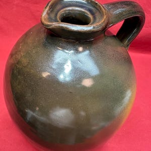 Pode incluir: Jarro de cerâmica verde escuro, feito à mão, com corpo arredondado e alça. O jarro apresenta uma ampla abertura circular e um acabamento brilhante, com variações de cor. Adequado para decoração de casa.