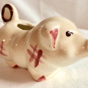 May include: A white ceramic pig creamer with pink and brown painted details. The pig has a spout on its snout and a handle on its back.