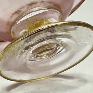 May include: Close-up of a clear glass footed dessert dish with a gold rim. The base of the dish is round and has a gold rim. The stem of the dish is clear glass with a gold accent. The bowl of the dish is pink.