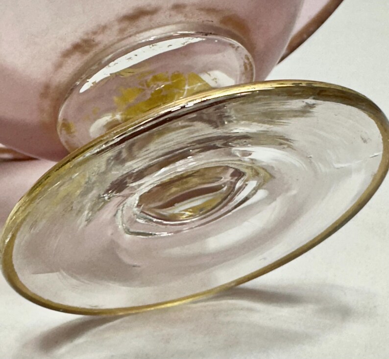 May include: Close-up of a clear glass footed dessert dish with a gold rim. The base of the dish is round and has a gold rim. The stem of the dish is clear glass with a gold accent. The bowl of the dish is pink.