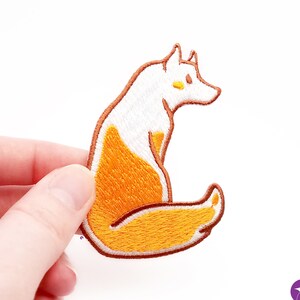 以下が含まれることがあります： 白い体とオレンジ色の尻尾を巻いた座っているキツネの刺繍パッチ