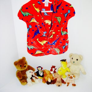 Puede incluir: Una camisa roja de manga corta con estampado de dinosaurios. La camisa tiene un frente abotonado y está colgada de una percha blanca. Hay varios animales de peluche sobre una superficie blanca debajo de la camisa.