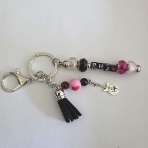 May include: A silver-toned metal keychain featuring a split ring and lobster clasp. One strand includes clear, black, and pink beads with letter cubes spelling PHJ and a heart icon. The second strand has a black tassel, pink and dark beads, and a guitar charm.