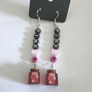 May include: Dangle earrings featuring silver-toned hooks, black letter beads spelling PINK, translucent light pink heart beads, small round pink beads with black hearts, and miniature red strawberry jam jar charms with labels.
