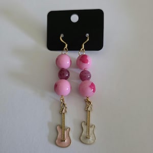 May include: A pair of dangle earrings featuring gold-tone hooks, each with two round pink beads, one translucent purple bead, and a small guitar-shaped charm at the bottom. The earrings are attached to a rectangular black display card with a hole.