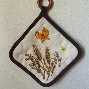 May include: A diamond-shaped fabric potholder with a dark brown border and a wooden hanging ring. The off-white center features a printed illustration of brown wheat stalks, a large orange butterfly, and a small yellow butterfly with the text Franco.
