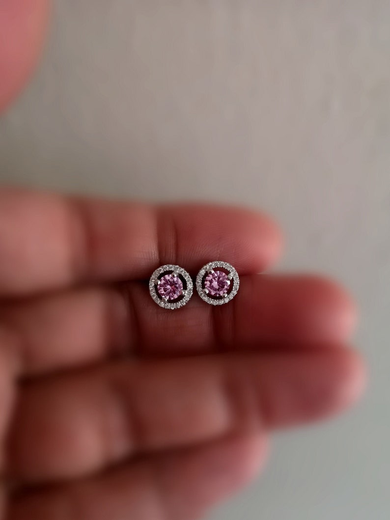 May include: A pair of silver stud earrings with a pink gemstone surrounded by a halo of smaller clear gemstones.