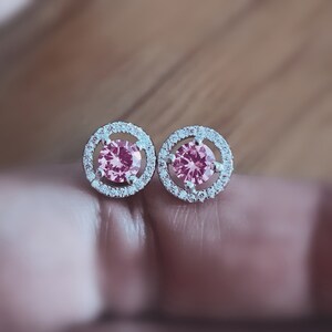 May include: A pair of silver earrings with a pink gemstone surrounded by a halo of smaller clear gemstones.