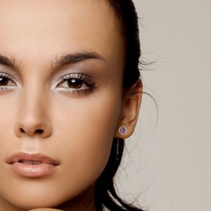 May include: A close-up of a woman's face with a small pink gemstone earring in her right ear. The earring has a halo of smaller clear gemstones.