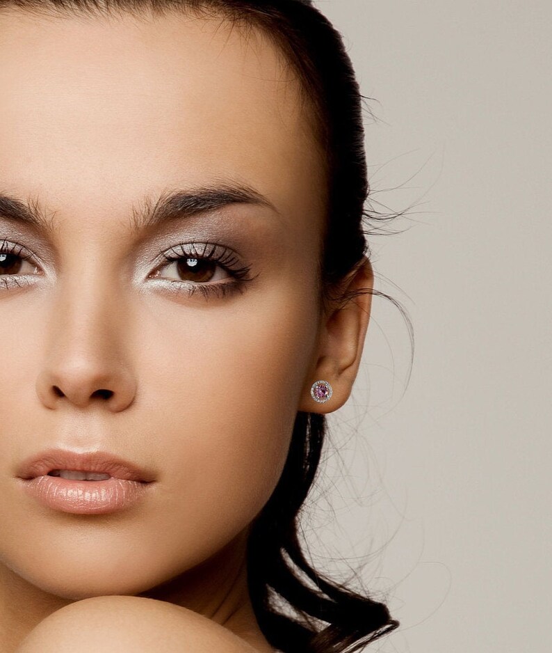 May include: A close-up of a woman's face with a small pink gemstone earring in her right ear. The earring has a halo of smaller clear gemstones.