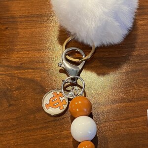 Puede incluir: Un llavero de pompón blanco y esponjoso con un cierre plateado y un dije con un logotipo naranja. El llavero también tiene una cadena de cuentas marrones y blancas alternadas.