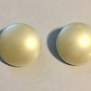 Puede incluir: Dos pendientes redondos de color blanco roto con acabado nacarado. Los pendientes tienen una forma ligeramente abovedada y una superficie lisa, que refleja la luz. Se presentan sobre un fondo liso de color claro.