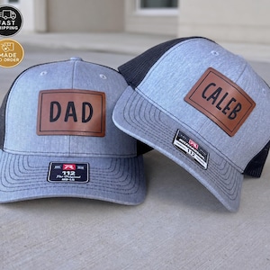 May include: Two heather grey and black trucker hats with mesh backs. Each hat features a rectangular brown leather patch on the front. One patch is engraved with the text DAD, and the other patch is engraved with the text CALEB. Both hats have a black sticker on the brim.