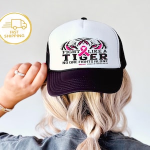 May include: A black and white trucker hat with the text "FIGHT LIKE A TIGER" and a pink ribbon, symbolizing breast cancer awareness. The hat features a tiger graphic and the phrase "NO ONE FIGHTS ALONE".