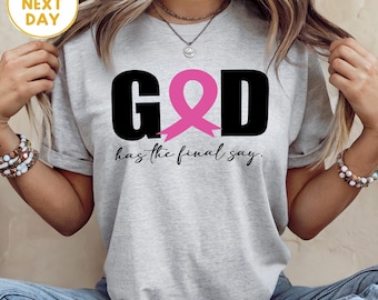 May include: A light gray t-shirt with the text "GOD" in black, featuring a pink ribbon in place of the "O". Below the text is the phrase "has the final say." The shirt is worn with blue jeans.