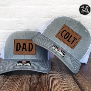 May include: Two heather gray and white trucker hats featuring rectangular brown leather patches. One patch is engraved with the word DAD and the other with the name COLT. Each hat has a black Richardson 112 sticker on the brim and white mesh back.