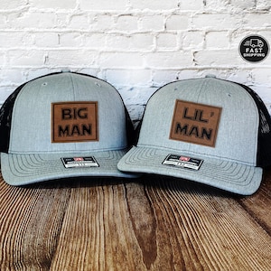 May include: Two gray trucker hats with black mesh backs. Each hat has a brown leather patch with the words "BIG MAN" and "LIL' MAN" in bold letters. The hats are on a wooden surface.