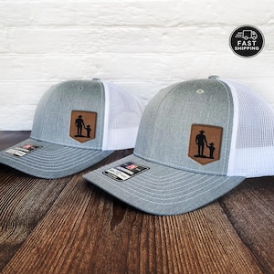 May include: Two gray trucker hats with white mesh backs. Each hat features a brown leather patch with a silhouette of a person and child. The hats have a curved brim and a Richardson brand tag.