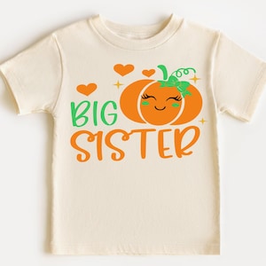 May include: A cream-colored short-sleeve t-shirt featuring a graphic of an orange pumpkin with a green bow, eyelashes, and a smile. The text reads BIG SISTER in green and orange, accompanied by orange hearts and small four-pointed star accents.