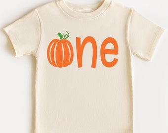 May include: A cream-colored short-sleeve t-shirt featuring a graphic print on the front. The design includes an orange pumpkin with a green stem and the lowercase word one in orange lettering, where the letter o is replaced by the pumpkin.