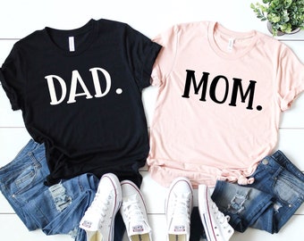 Peut inclure: Deux t-shirts, un noir avec "DAD." en texte blanc et un rose avec "MOM." en texte noir. Les t-shirts sont disposés sur une surface en bois blanc avec deux paires de baskets blanches et deux paires de jeans bleus.