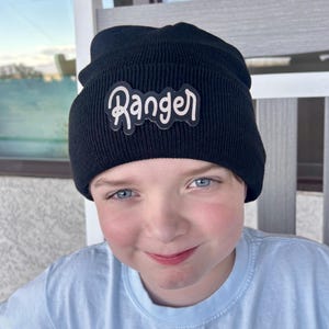 May include: A black knit beanie featuring a patch on the front with the word Ranger written in a stylized, light-colored cursive font. The hat has a ribbed texture and a folded cuff design, providing a snug fit around the head.